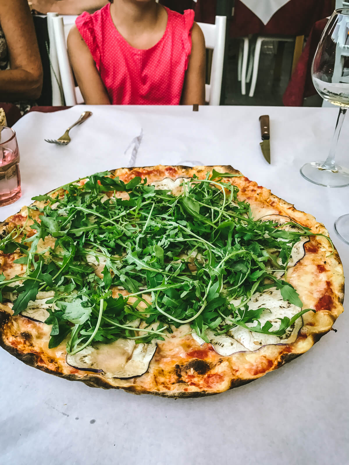 A large thin crust pizza with pizza sauce, cheese, eggplant and arugula on a white table in Tuscany Italy with a young girl in the background. 