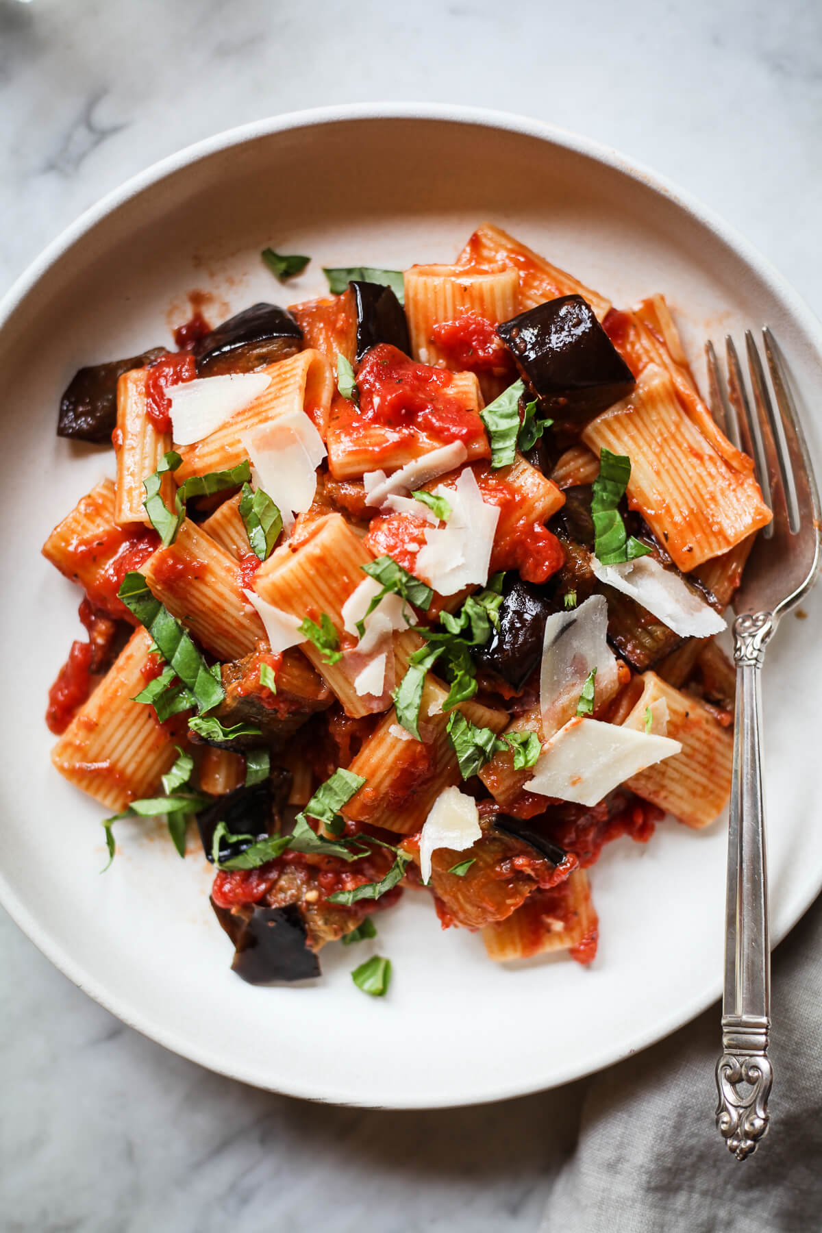 A white ceramic bowl filled with pasta alla norma - an eggplant and tomato ragu served with rigatoni and topped with basil. 