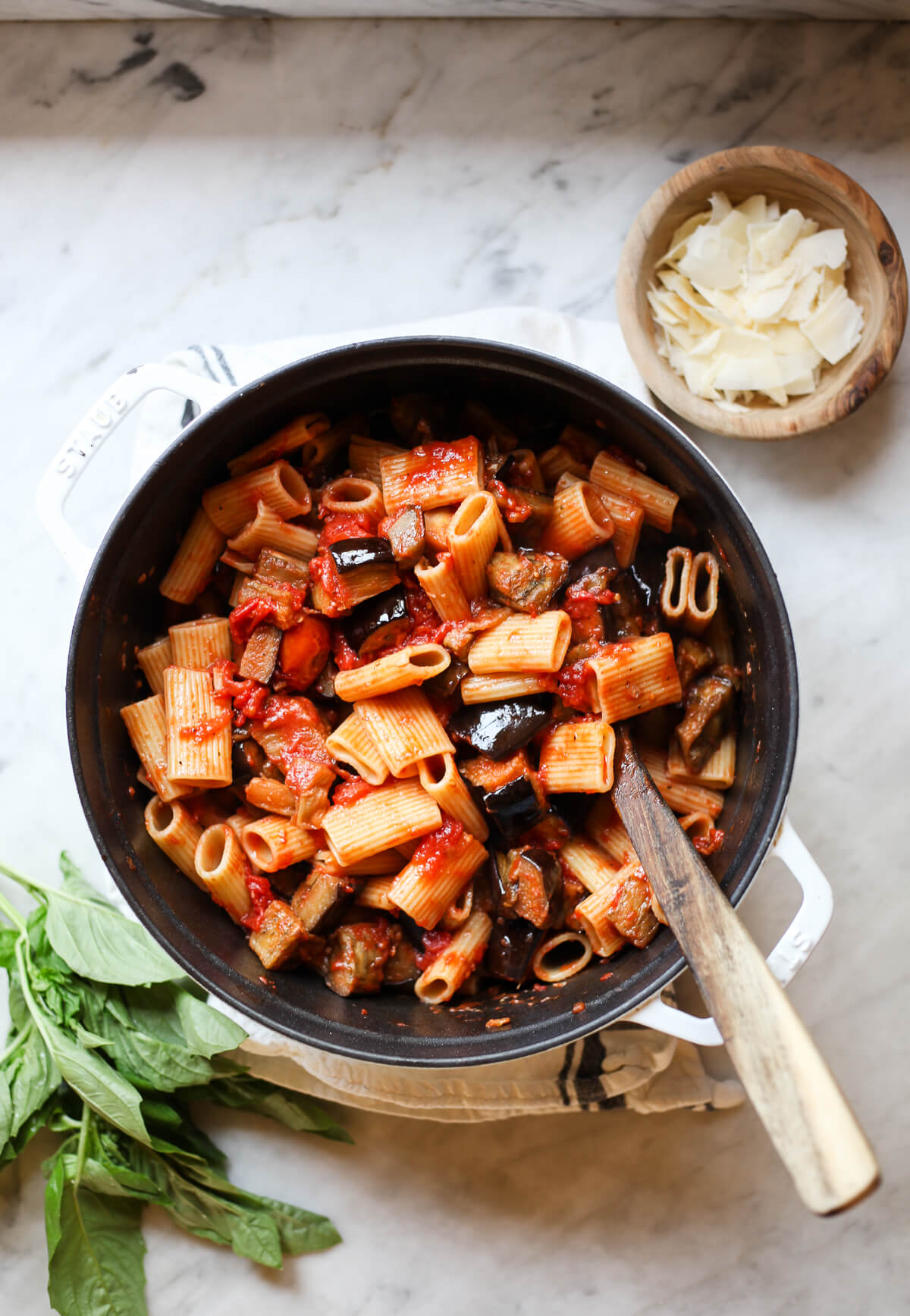 Pasta alla Norma Recipe A white Staub pot filled with Sicilian pasta alla norma recipe with eggplant ragu and tomatoes. The pot sits on a marble countertop with fresh basil and Parmesan cheese on the side.