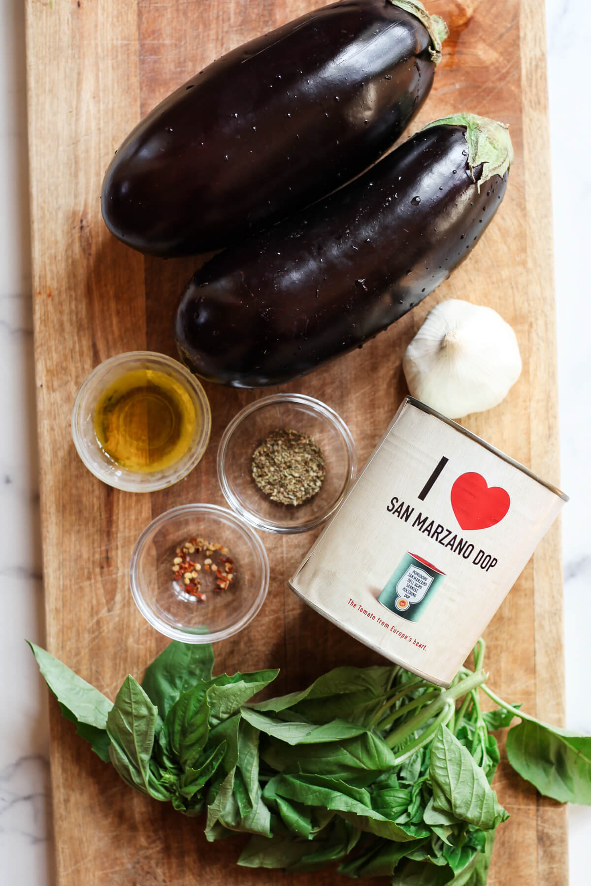 pasta alla norma ingredients Two eggplants, a can of san marzano dop tomatoes, olive oil, garlic, and herbs on a wood cutting board.