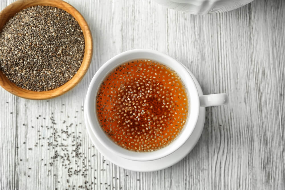 A white mug filled with herbal tea and chia sees on a white wooden counter next to a small bowl of chia seeds. 