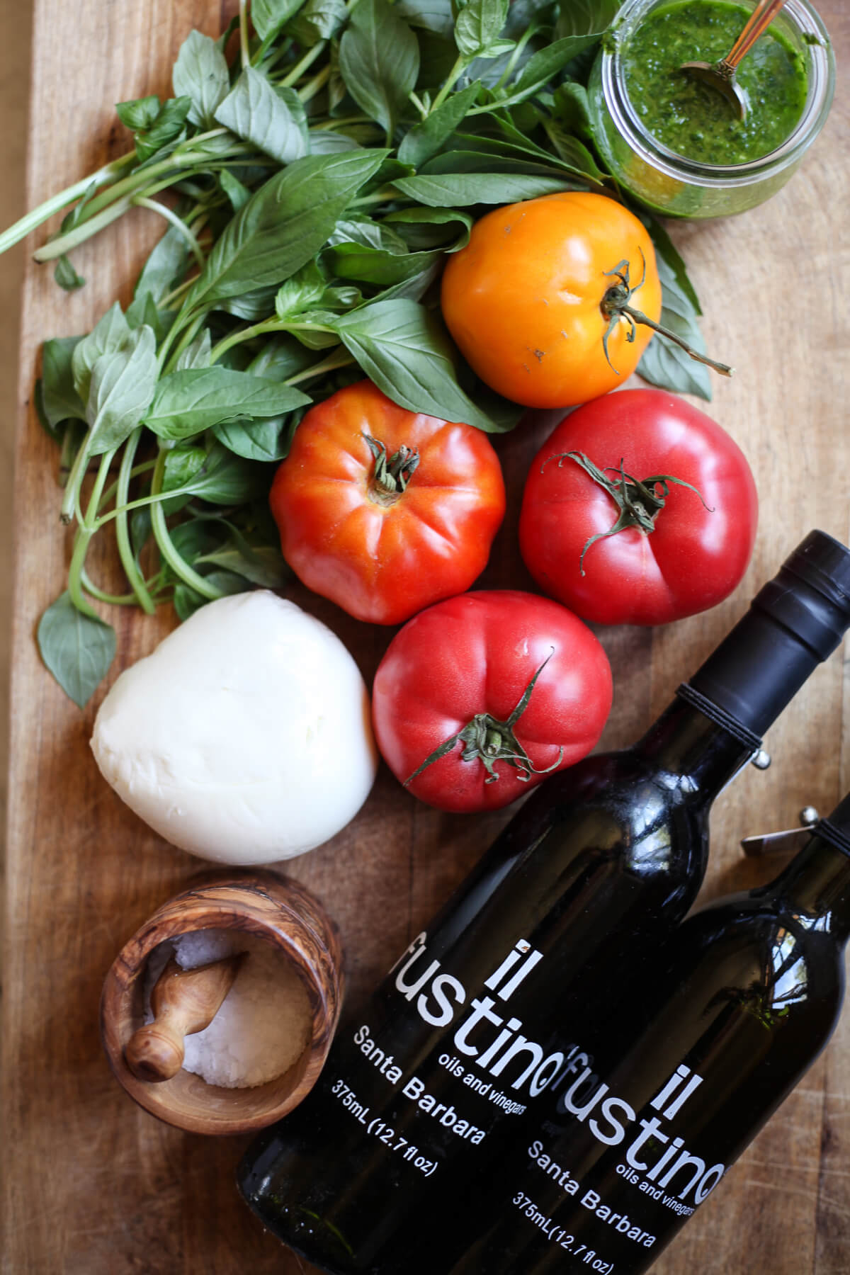Fresh basil, tomatoes, a ball of mozzarella, olive oil bottles, sea salt, and basil oil on a wooden cutting board. 
