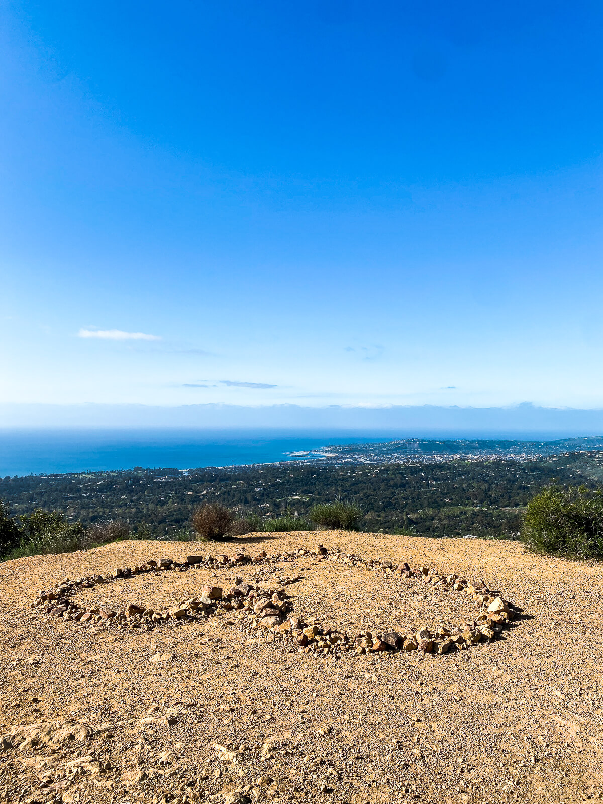 The view from the top of a mountain during a hike in Santa Barbara. A heart is made out of rocks on the ground and the ocean is seen in the background. 