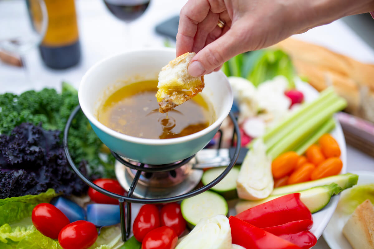 A woman's hand dips a piece of bread into a small bowl of bagna cauda that sits above a platter of vegetables.