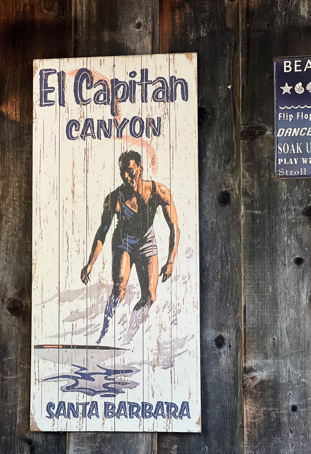 A vintage-inspired wooden sign with a painting of a surfer and text that reads "El Capitan canyon Santa Barbara" 