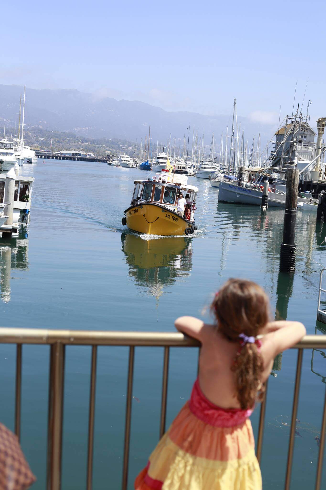 A cute yellow boat in the Santa Barbara harbor approaches a little girl wearing a bright orange, pink, and yellow dress. 