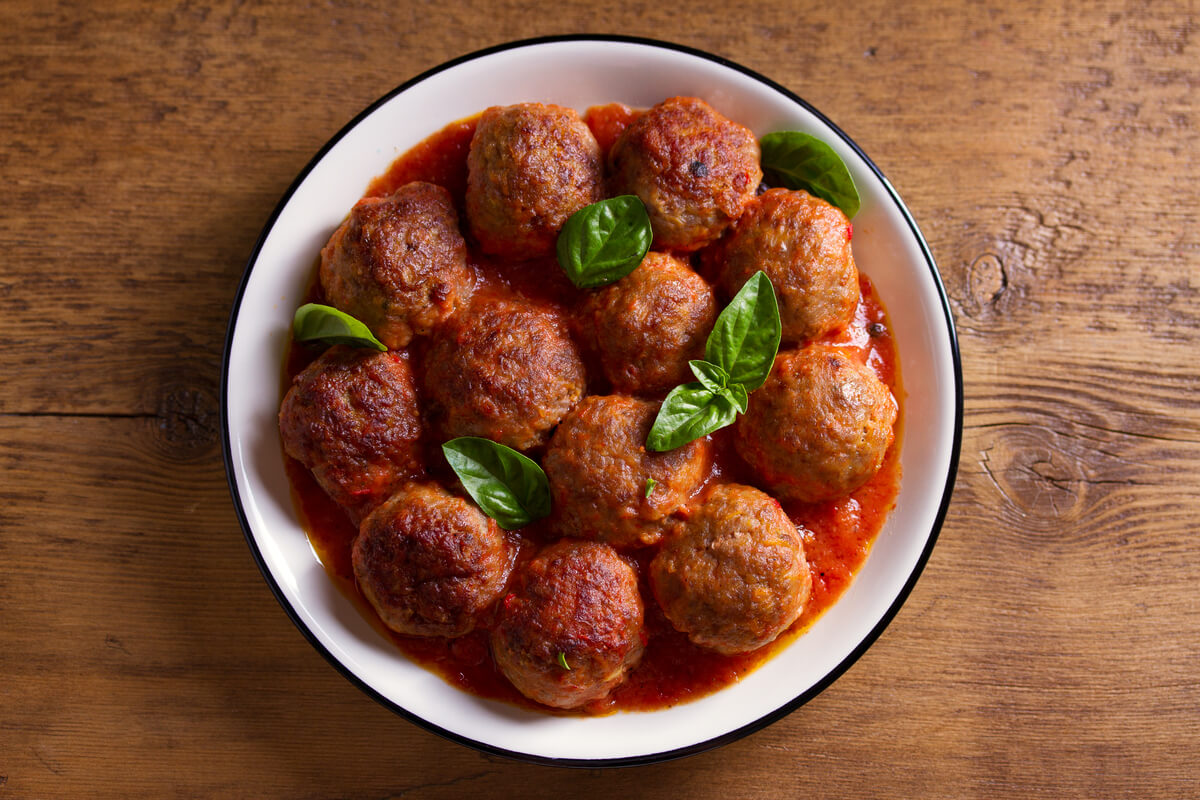 A white bowl filled with Italian meatballs in tomato sauce and garnished with basil leaves. The bowl sits on a wooden table. 