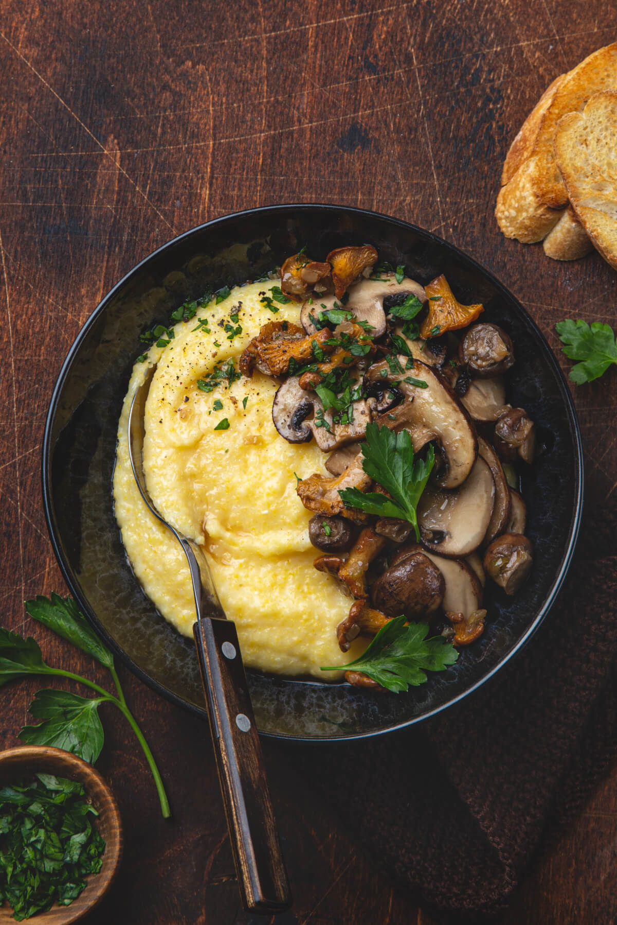 A dark bowl filled with creamy polenta topped with mushrooms and parsley sits on a dark wooden counter. 