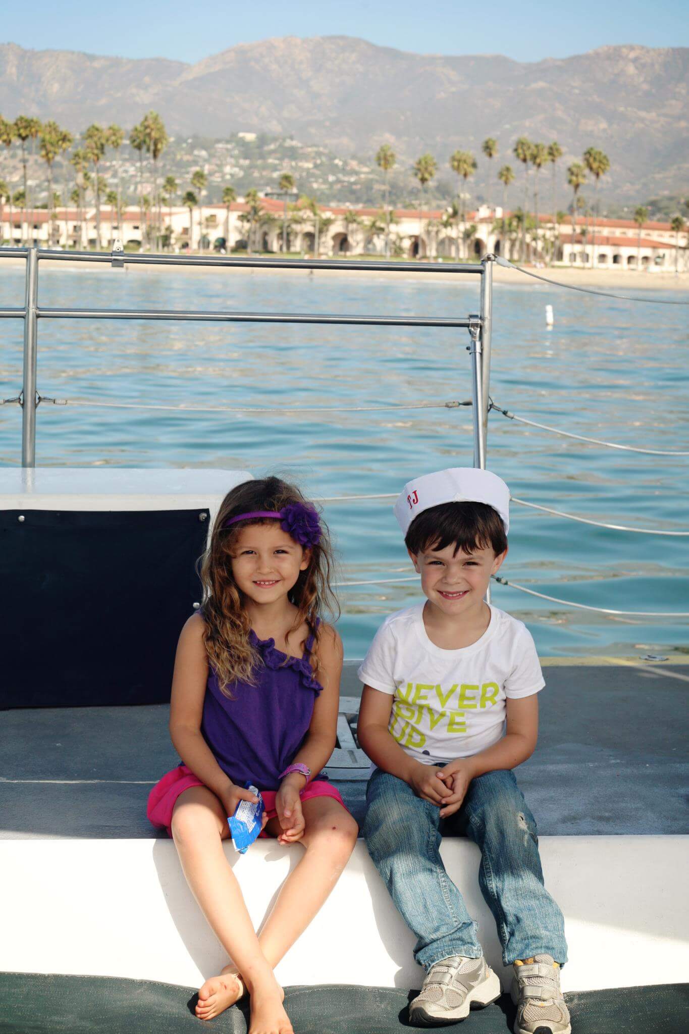 A little girl and boy sit on a boat in the ocean with Santa Barbara palm trees, buildings, and mountains in the background. This is an example of fun things to do in Santa Barbara with kids. 