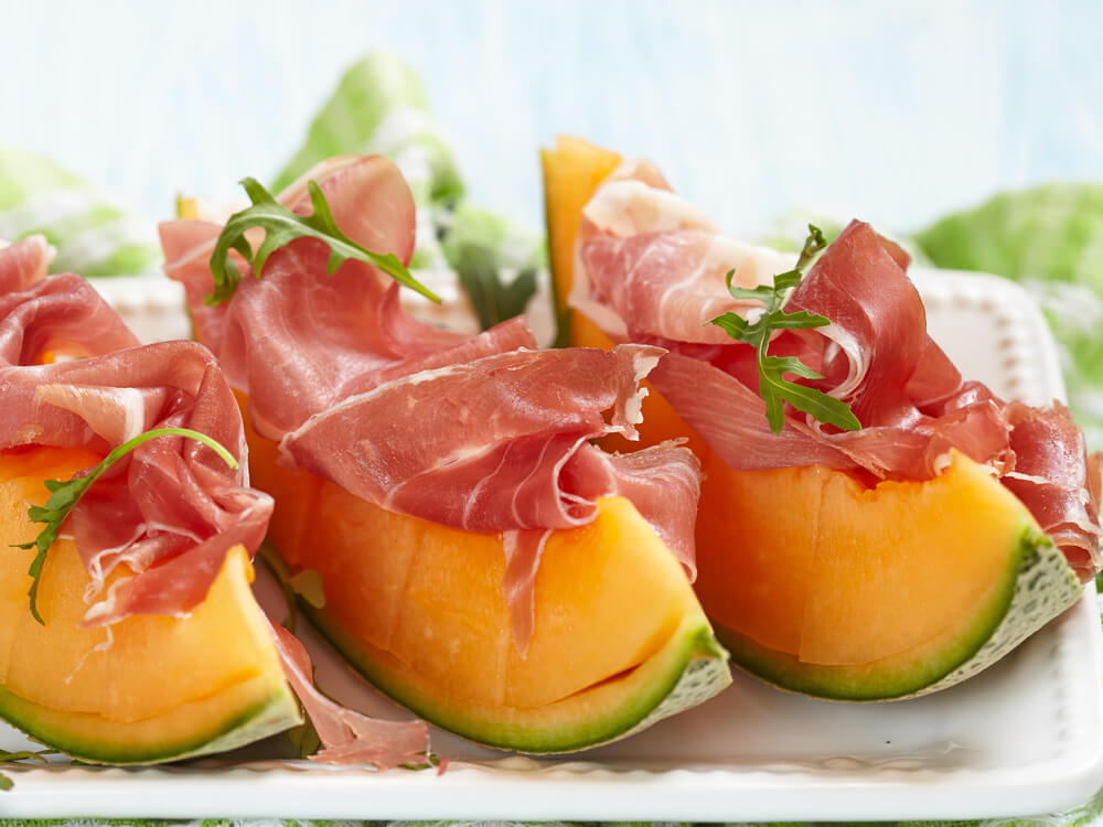 Three wedges of cantaloupe topped with slices of prosciutto and arugula for a traditional Italian food served as an antipasto. 