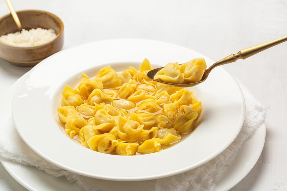 Tortellini in brodo (tortellini in broth) in a white bowl with a spoon lifting two tortellini out. A small bowl of cheese in the background. 