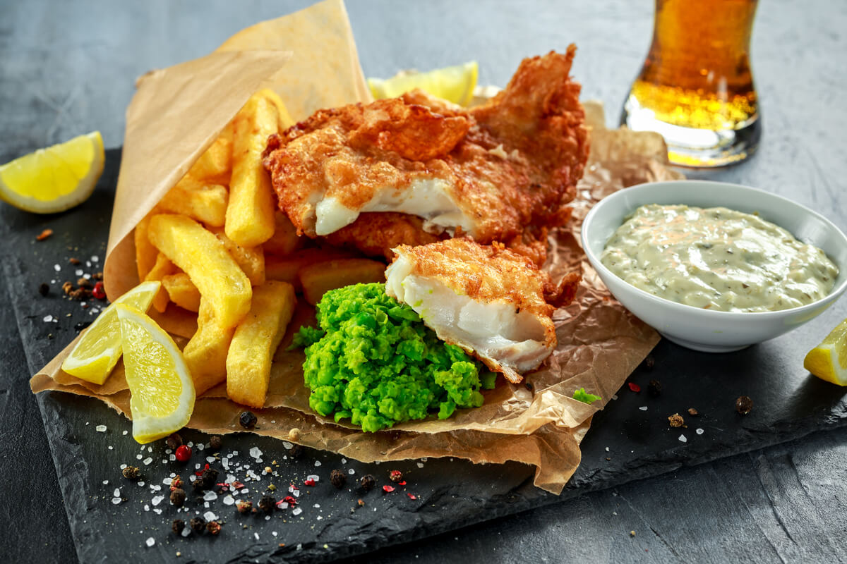 A traditional British fish and chips dish on parchment paper served with a side of mushy peas and tartar sauce. 