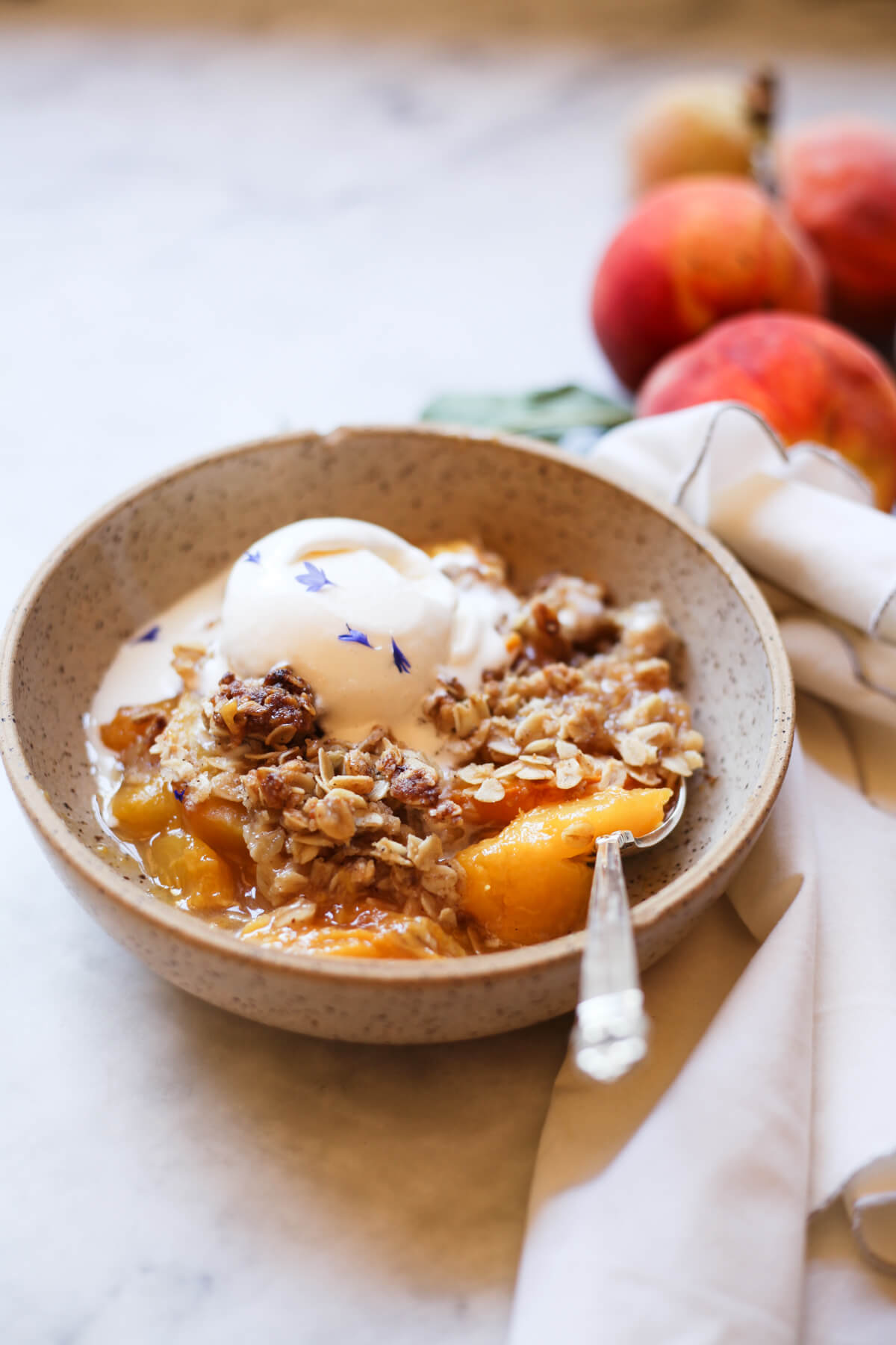 Peach crisp with oat crumble topping and a scoop of vanilla ice cream in a earthenware bowl on a white marble countertop. 