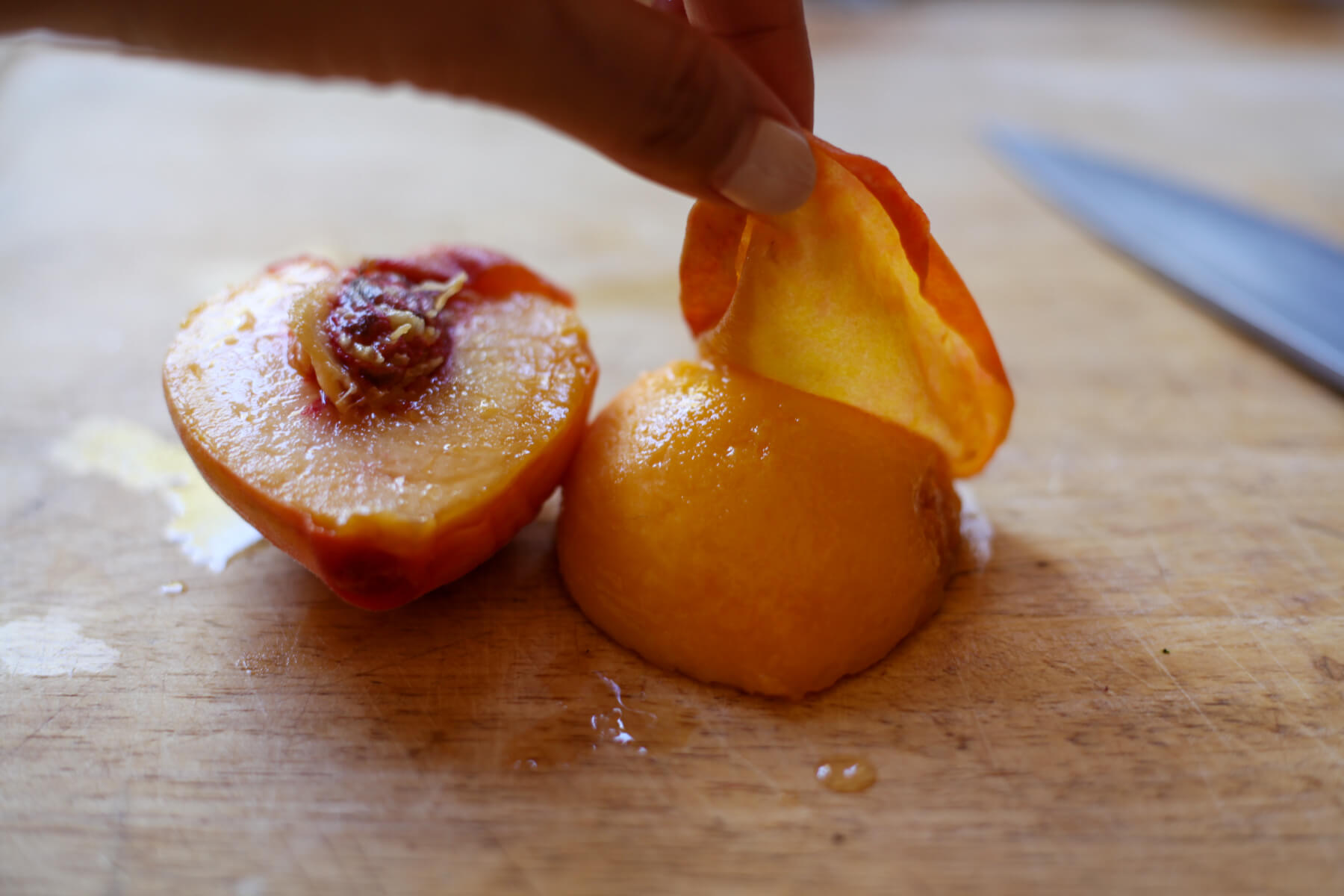 A peach is halved on a cutting board and a hand peels off the skin of one half.