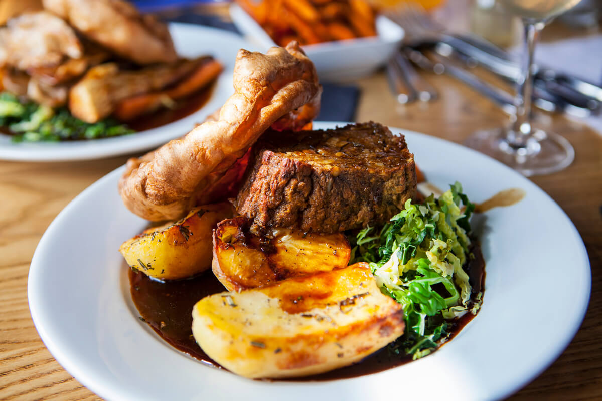 A piece of plant-based meat roast over roasted potatoes with a Yorkshire pudding and greens on a white plate. A traditional British food. 