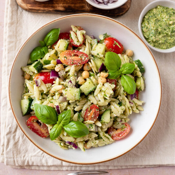 A white bowl filled with pesto orzo pasta salad with cherry tomatoes, basil, cucumber, and chickpeas.