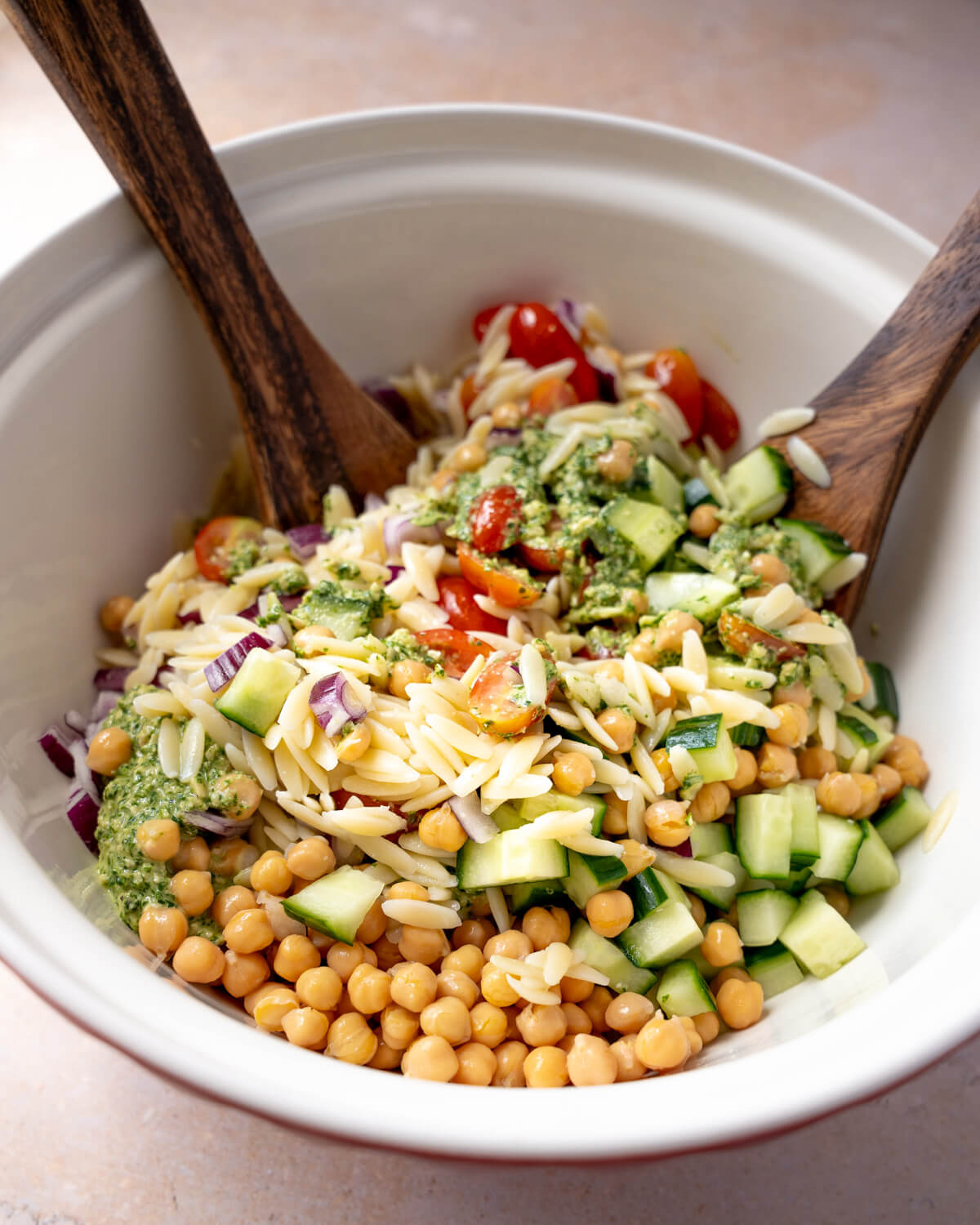 Cooked orzo, chickpeas, cucumber, tomatoes, red onion, and pesto are tossed together with wooden spoons in a white salad bowl to make a pesto orzo salad. 