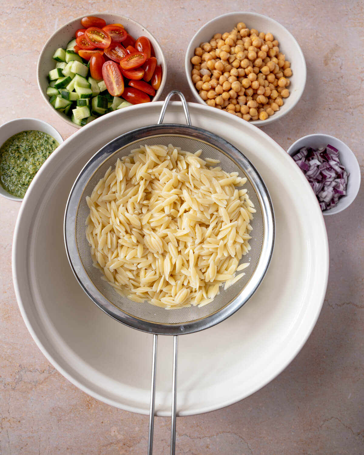 The ingredients for orzo salad around a white bowl. The orzo pasta salad ingredients include pesto, cucumber, tomatoes, chickpeas, and diced red onion..