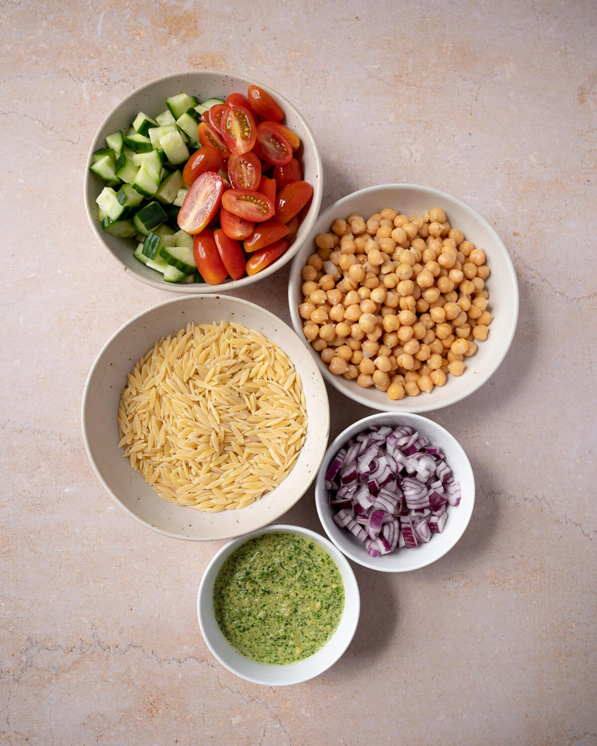 Bowls of the ingredients for making a pesto orzo salad. Clockwise from the top: cucumber and tomatoes, chickpeas, red onion, pesto, and dry orzo pasta. 