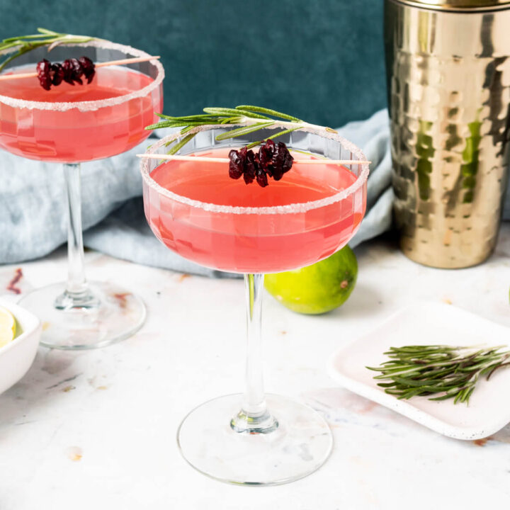 Two cranberry margaritas in coupe glasses garnished with dried cranberries and rosemary.