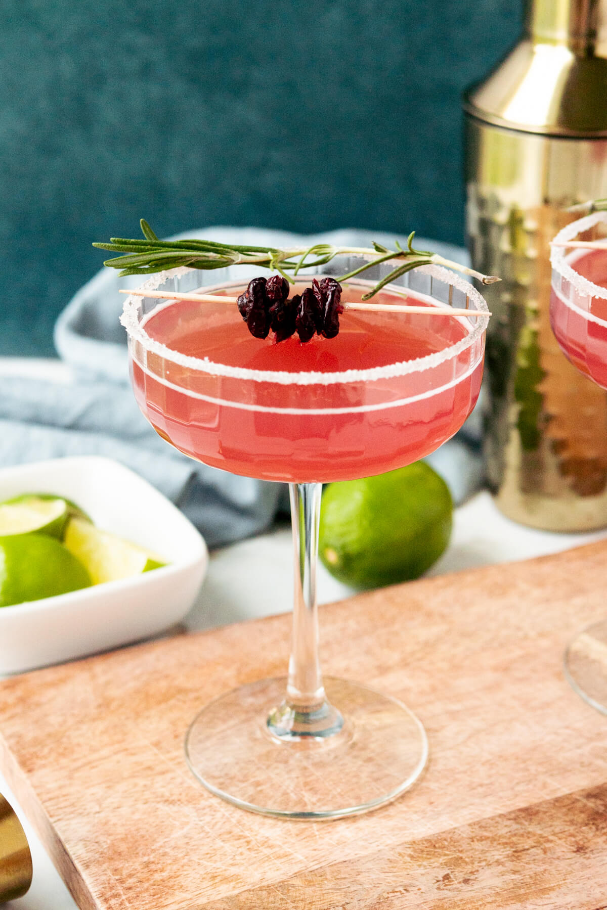 A cranberry Mistletoe margarita garnished with dried cranberries and rosemary in a coupe glass with limes and a cocktail shaker in the background. This is a delicious looking winter margarita recipe. 