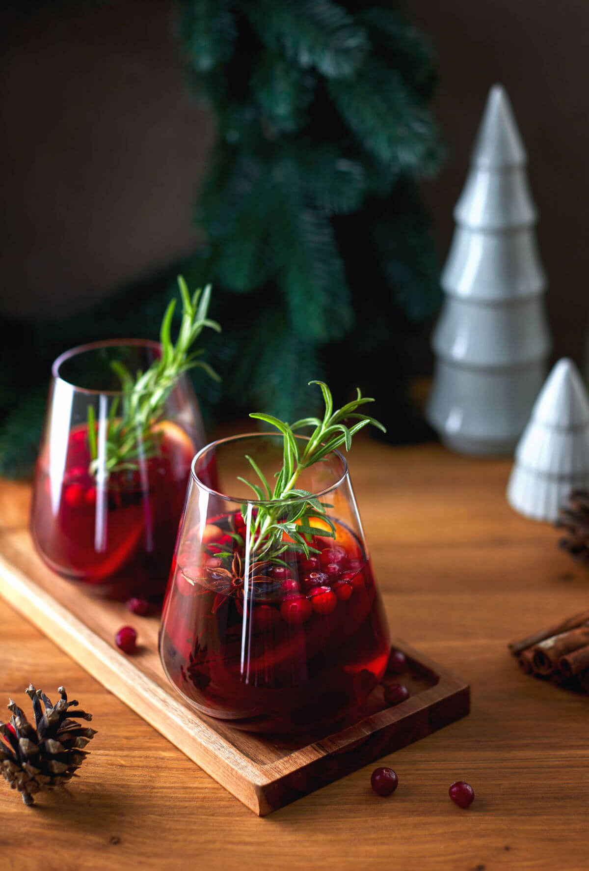 A beautiful photo of two stemless wine glasses filled with a holiday drink made with red wine, rosemary, orange, and cranberries. A wreath and silver Christmas tree decorations in the background. 