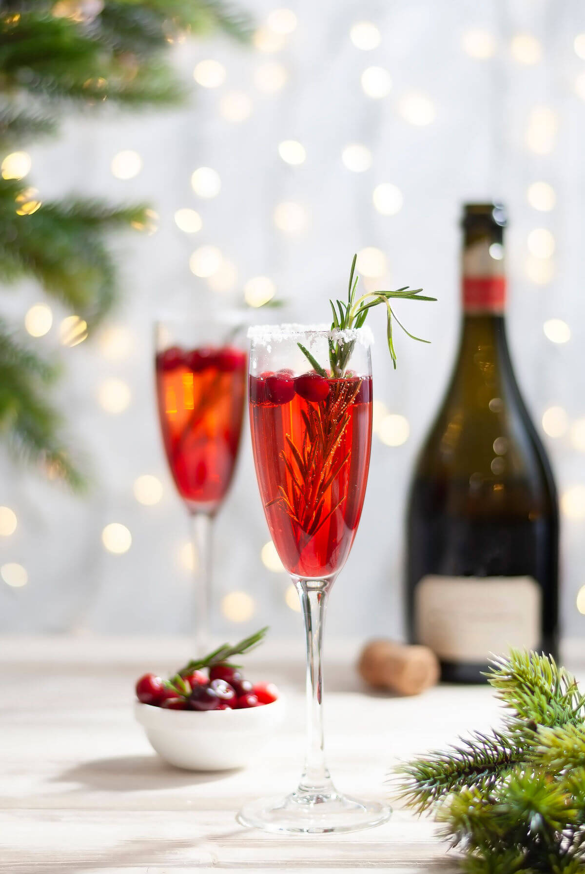 Two Poinsettia Cocktails made with Champagne and cranberry juice in front of a white background with christmas lights and a Champagne bottle. 