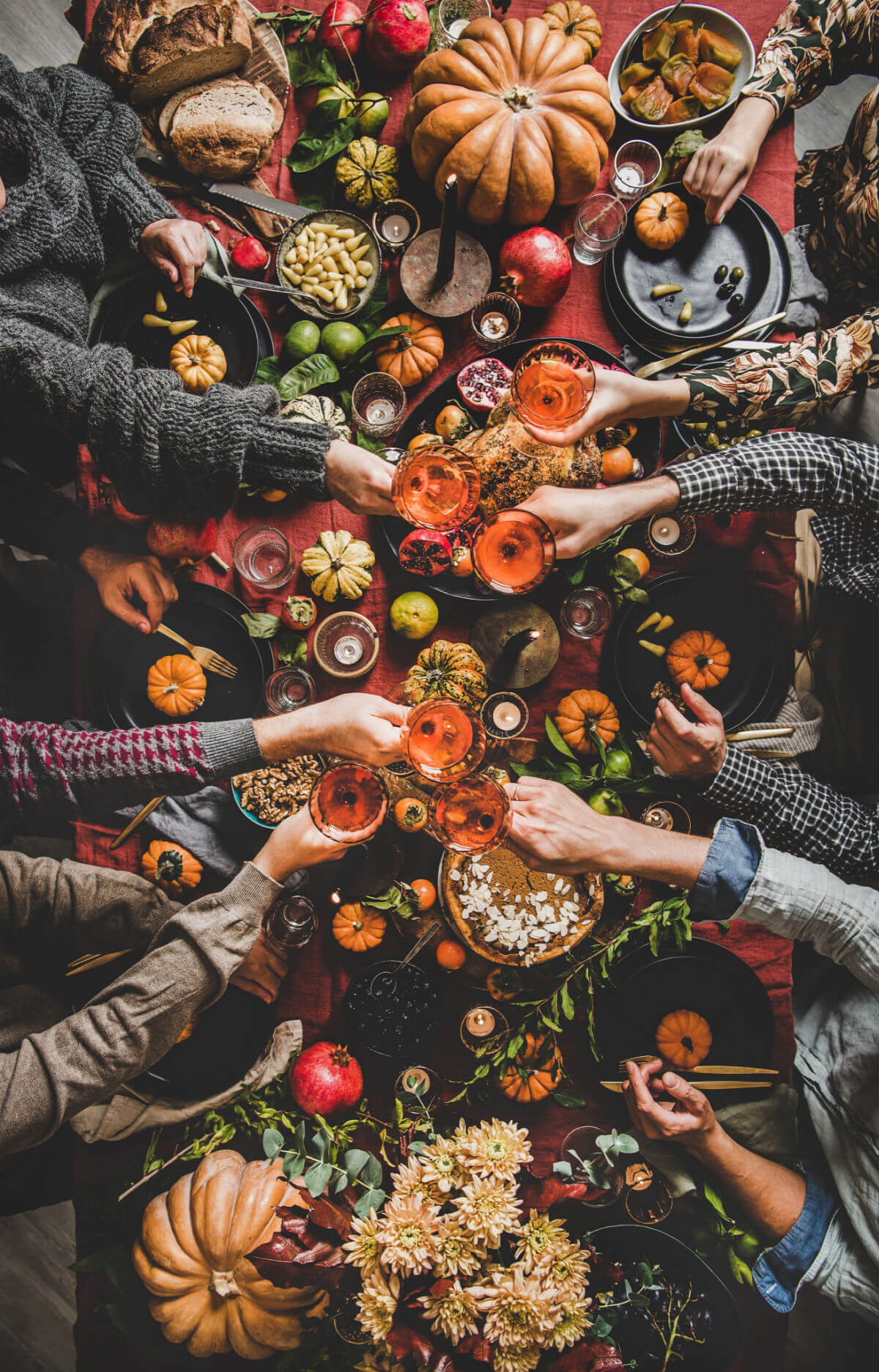 An overhead photo of people sitting raising wine glasses around a traditional holiday table with Thanksgiving colors including pumpkins and a red tablecloth. 
