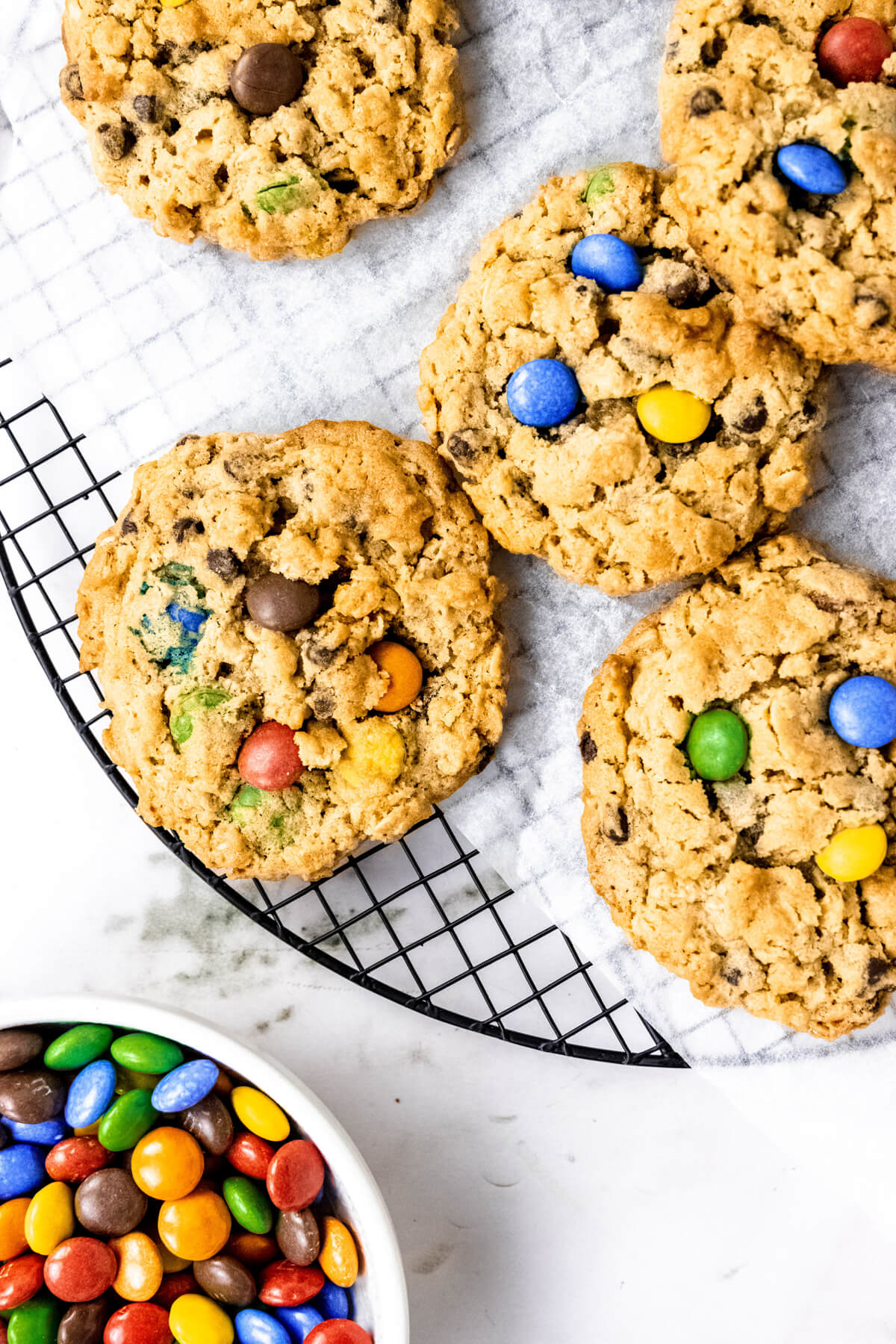 5 peanut butter monster cookies with oatmeal and M&Ms on a cooling rack with a bowl of m&ms on the side. 