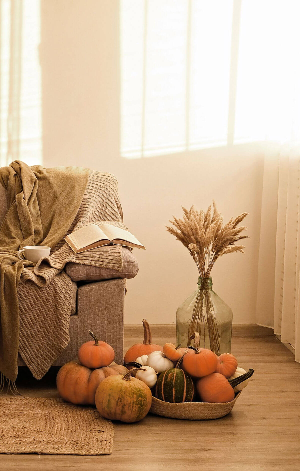 A serene living room space with a brown sofa, brown blankets, natural basket filled with pumpkins, and dried wheat in a glass vase on the floor. 