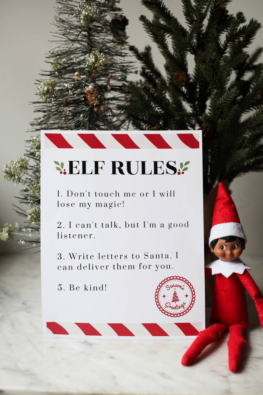 An Elf on the Shelf sits next two two small Christmas trees and a printable that says "Elf Rules" with a list of the Elf on the Shelf Rules. 