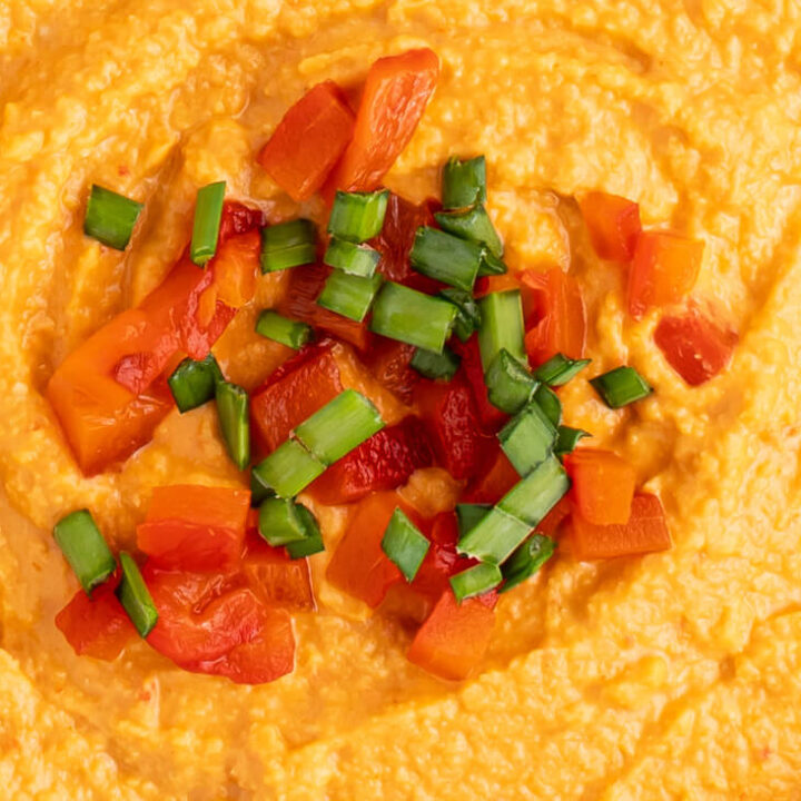 A close up photo of roasted red pepper hummus topped with chopped red peppers and green onions.