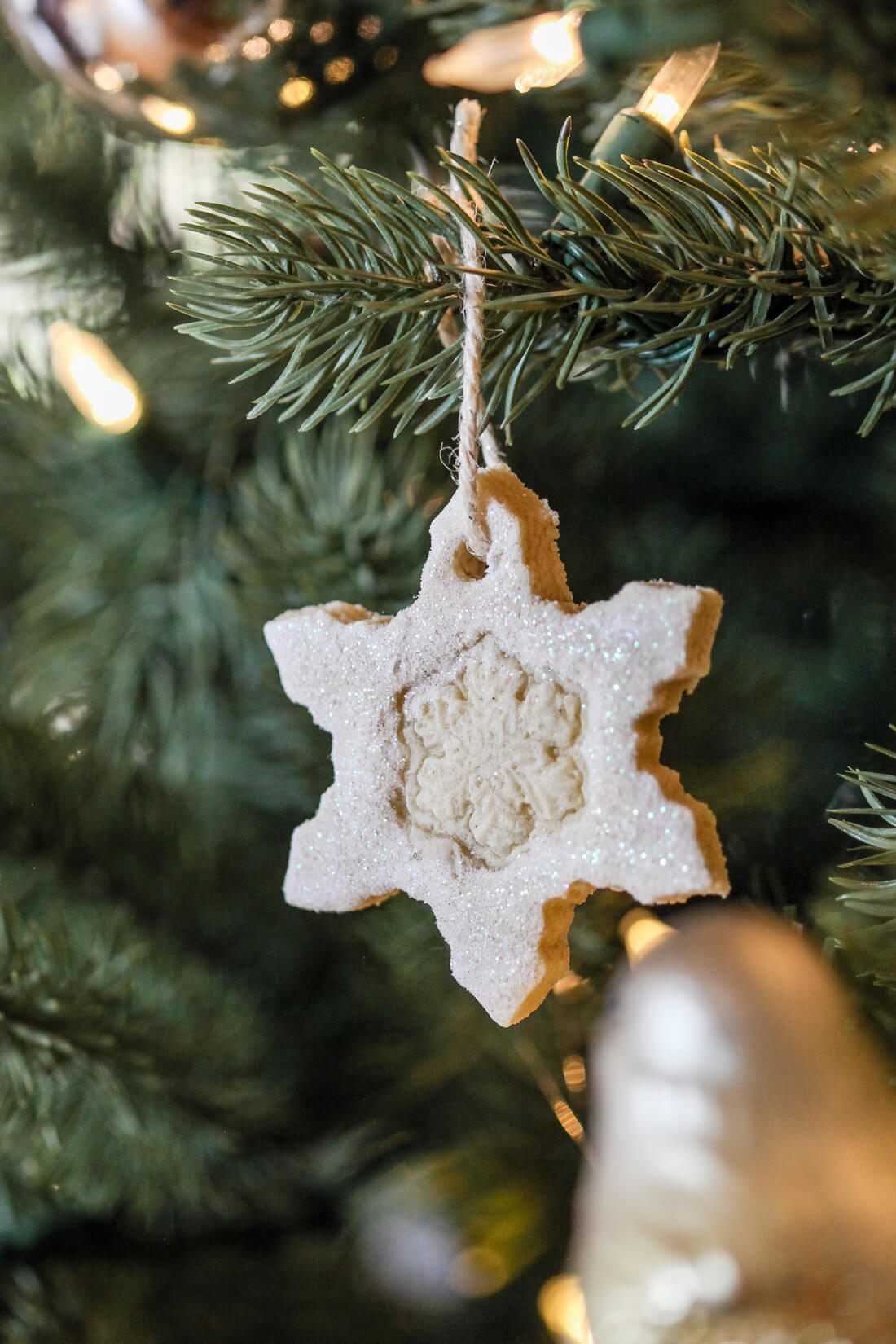 A white glitter coated salt dough snowflake ornament hangs from a Christmas tree branch by a pice of twine. 