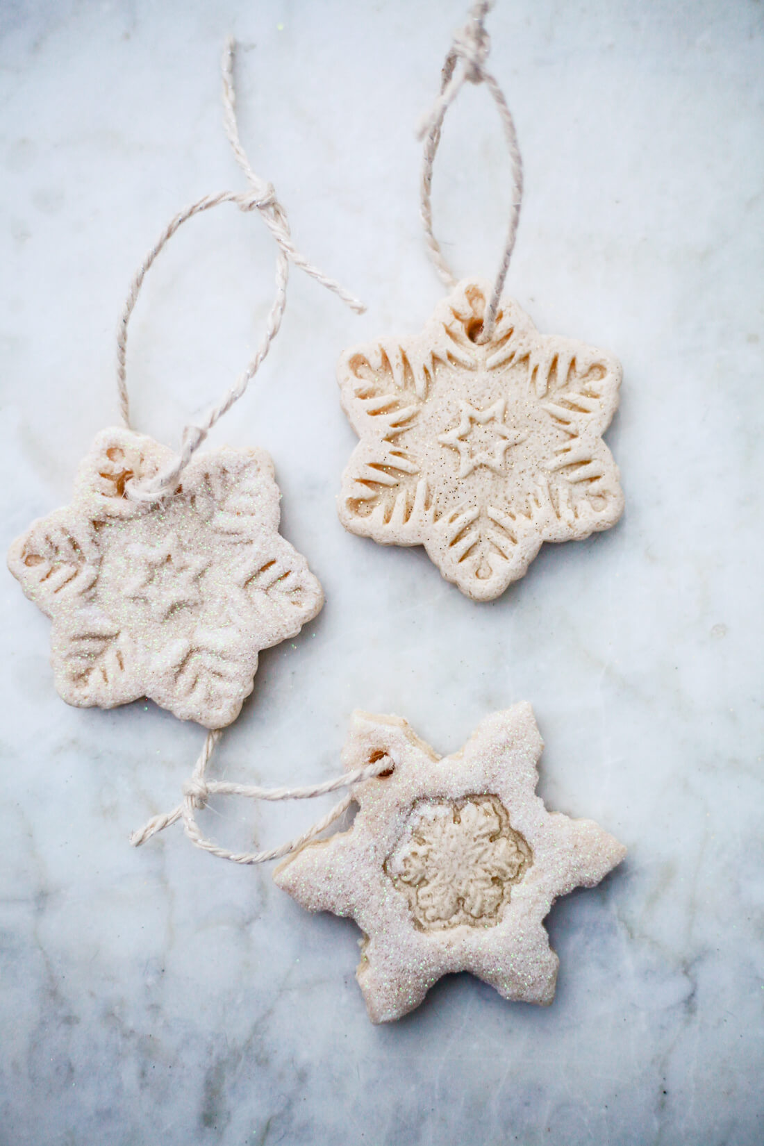 Three snowflake salt dough ornaments with twine and white glitter on a marble counter. 