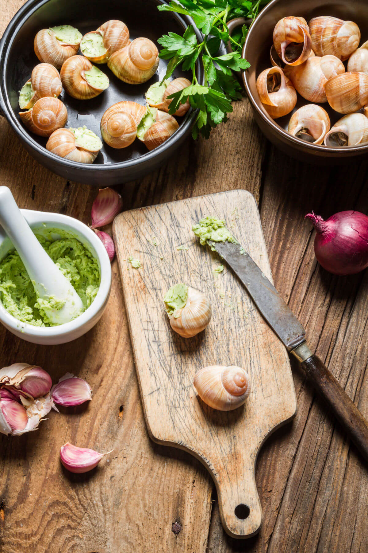 This photo shows how to make escargots de Bourgogne. There are bowls of snail shells, homemade parsley butter, and prepared escargots de Bourgogn on a wooden table. 