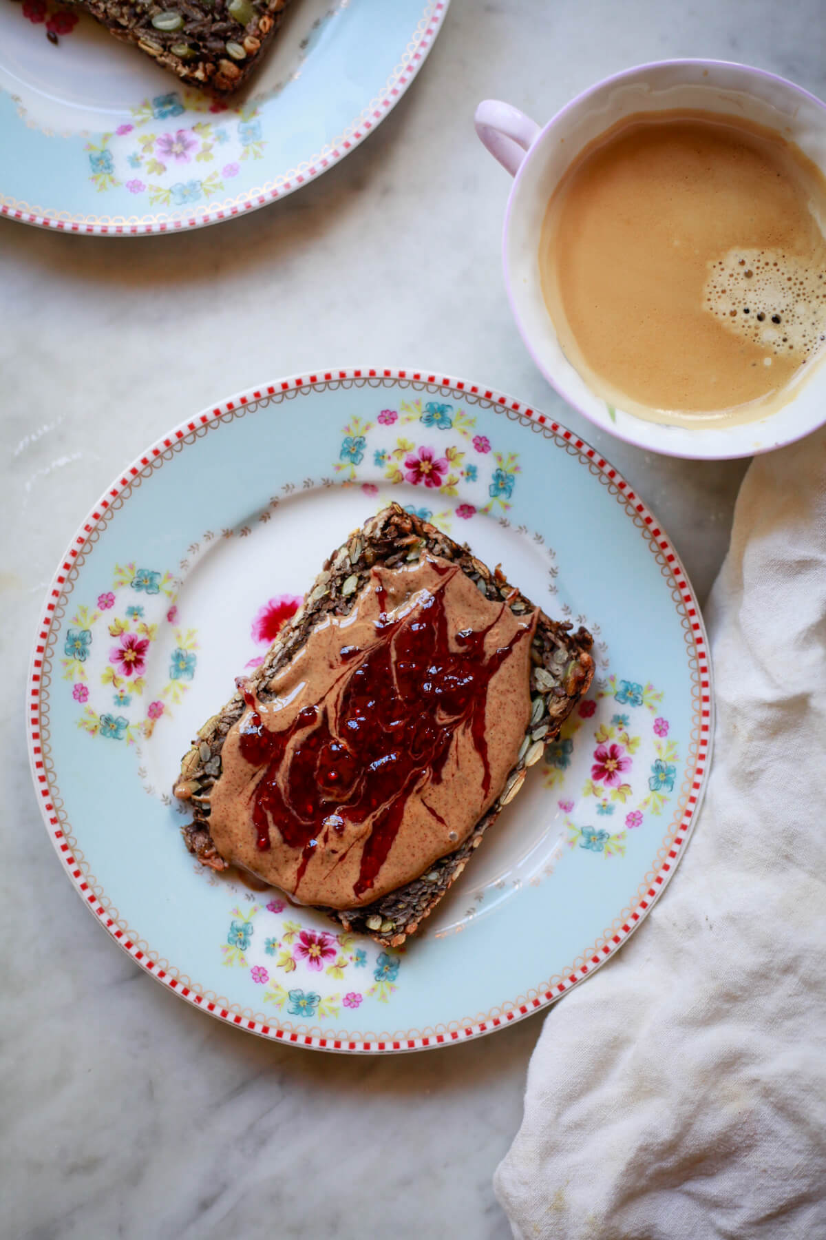 A slice of seed bread topped with peanut butter and jam on a floral plate next to a cup of coffee. A healthy gluten-free vegan breakfast made with seed bread. 