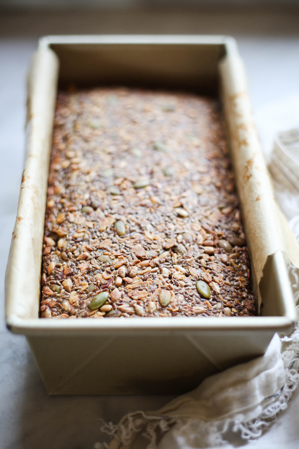 A freshly-baked loaf of super seed bread in a gold loaf pan lined with parchment paper on a white countertop. 