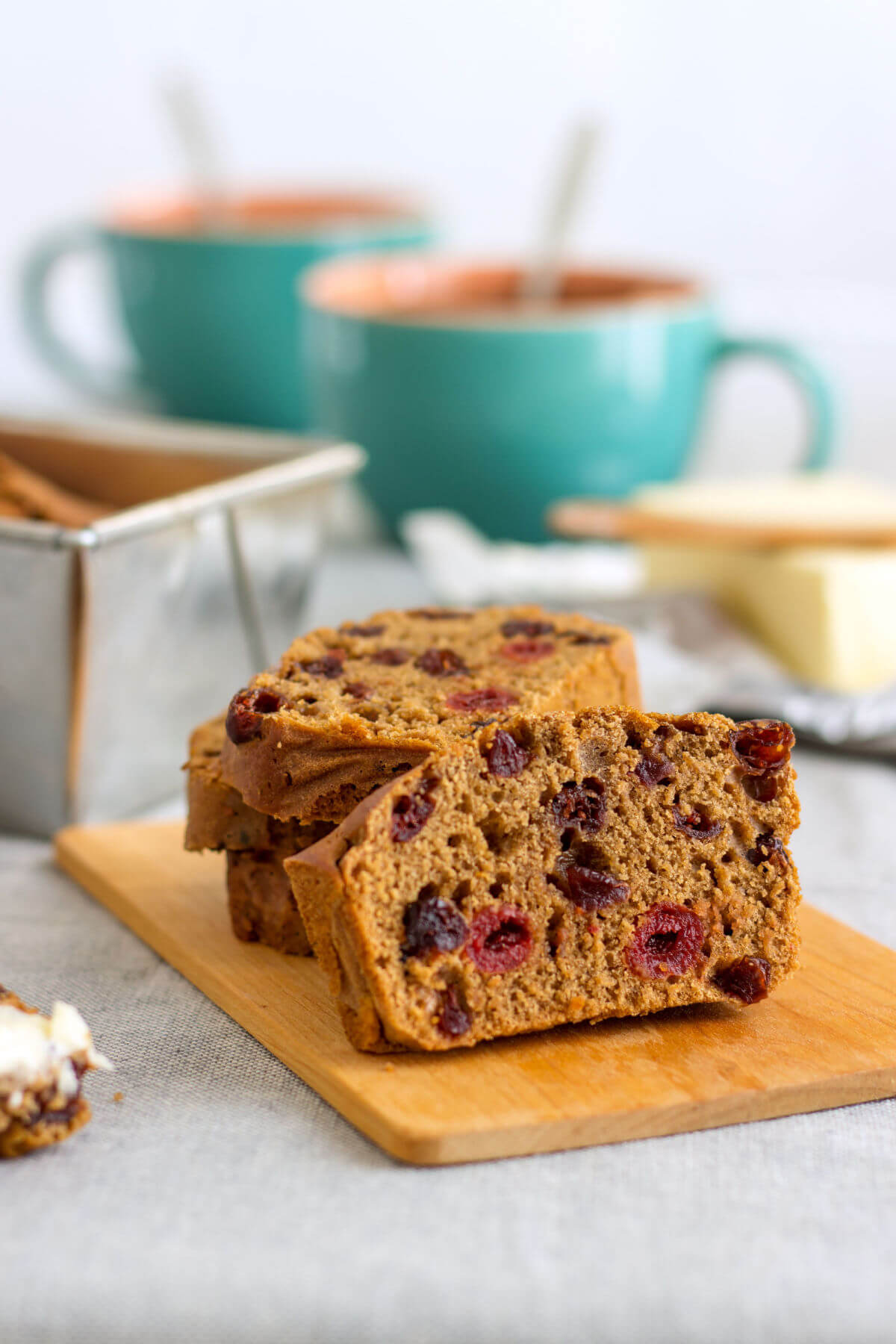 A loaf of homemade Irish barmbrack bread on a wooden cutting board with a loaf pan and coffee mugs in the background. This brown quickbread slice features cherries.