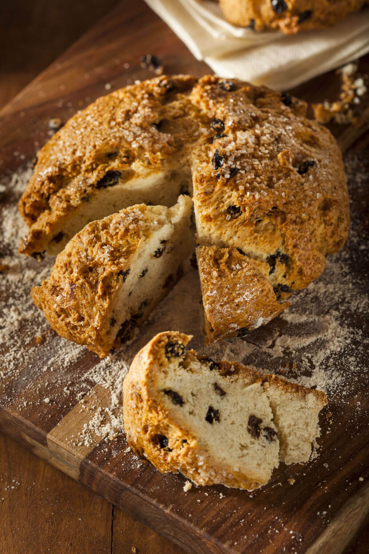 A homemade round loaf of Irish soda bread on a wooden cutting board. The bread is cut into wedges and made with raisins. 
