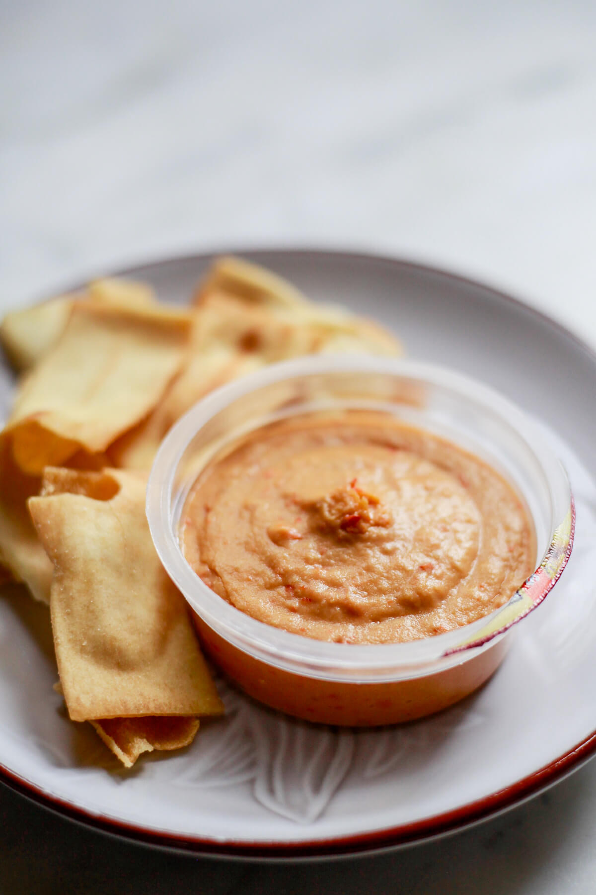 A Sabra red pepper hummus snack cup that was previously frozen on a white plate with pita chips. 