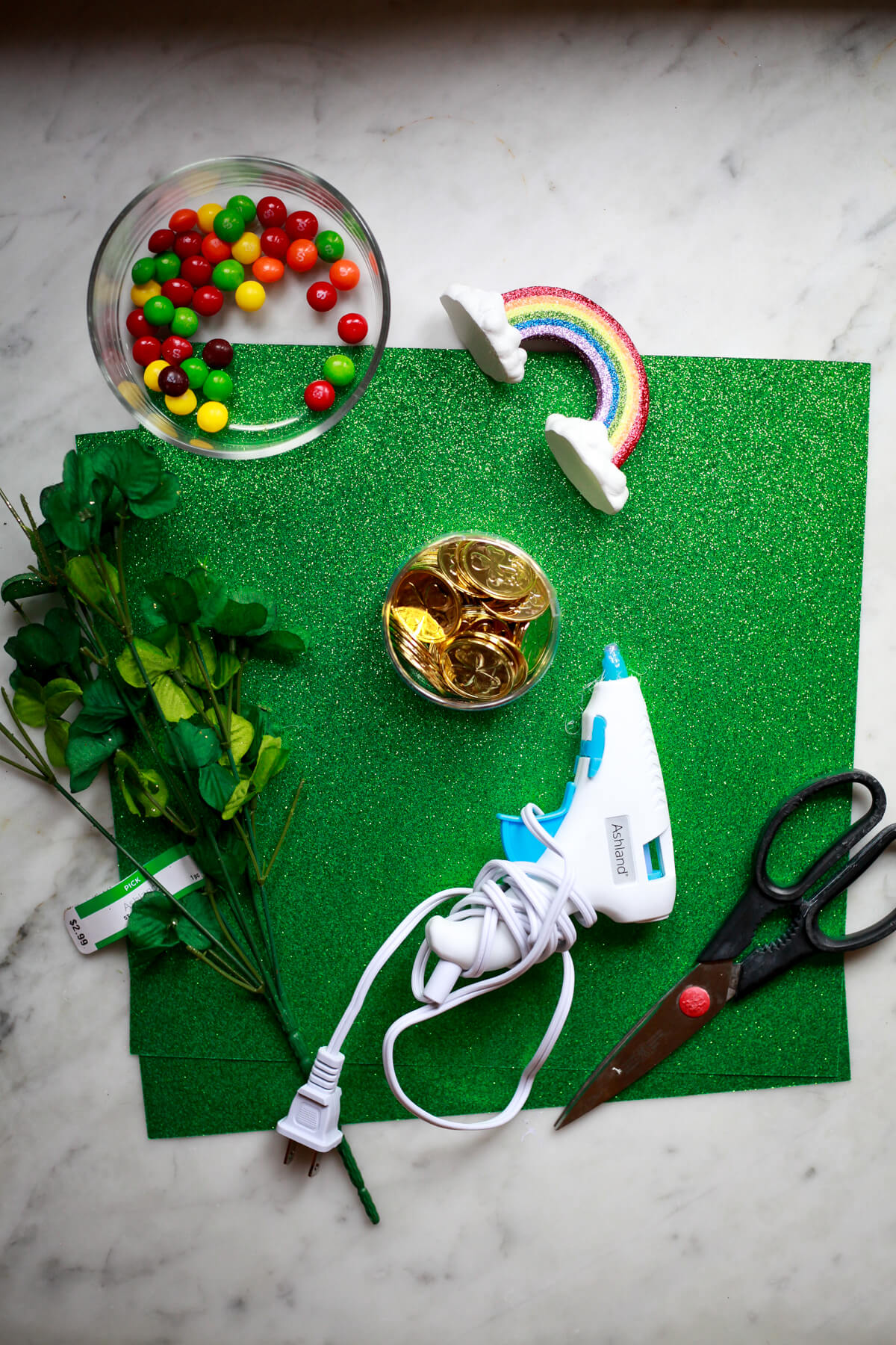 Green paper, skittles, a rainbow, gold coins, faux cloers, a glue gun, and scissors on a marble countertop to show leprechaun trap supplies. 
