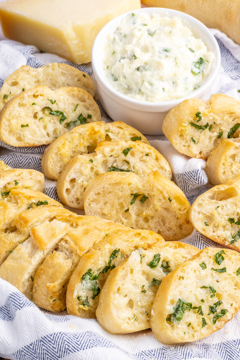 Slices of garlic bread on a kitchen towel with a small white dish of garlic herb butter in the background. 