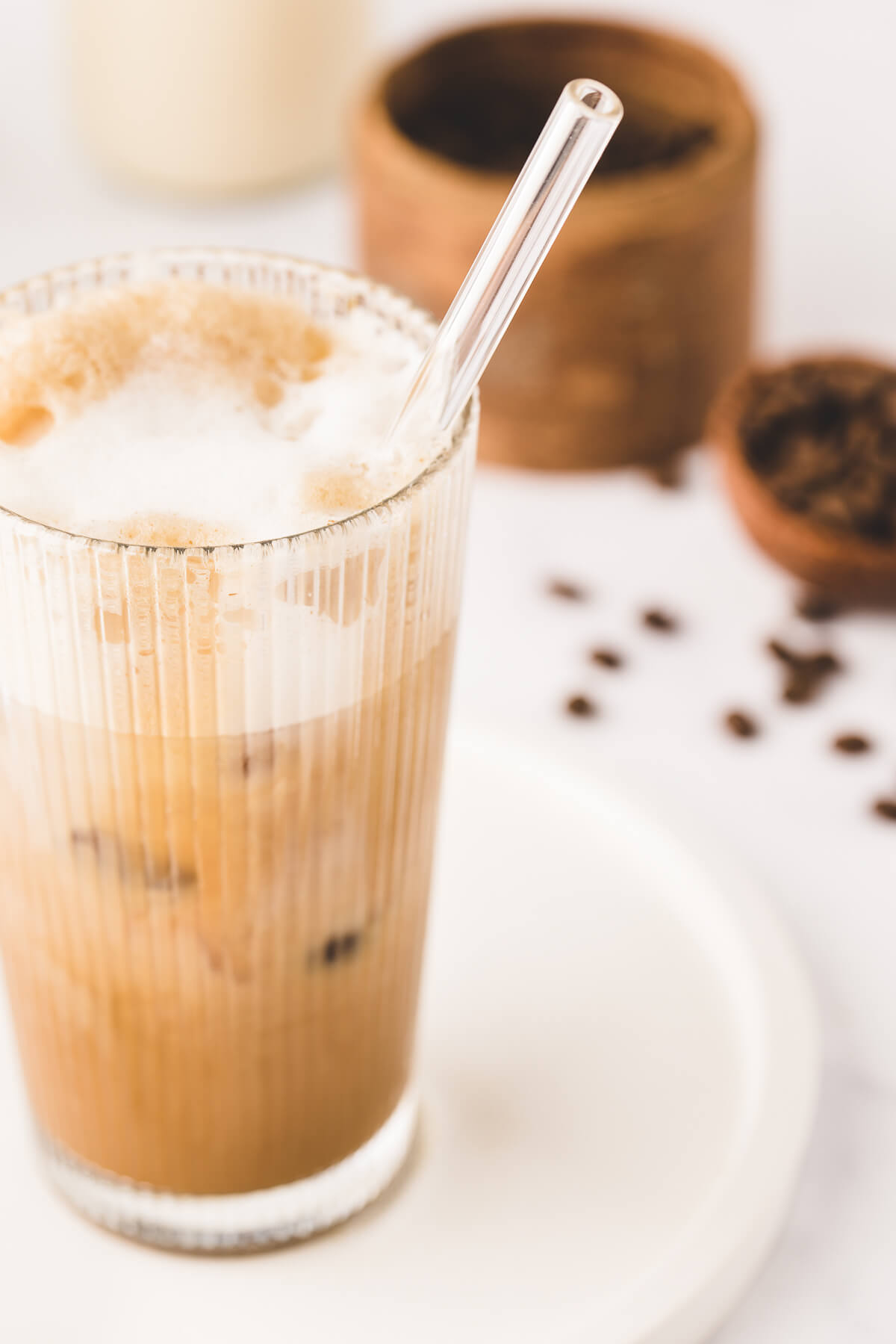 An oatmilk iced shaken espresso with cold foam in a tall glass with glass straw. Coffee beans in the background on a white counter. 