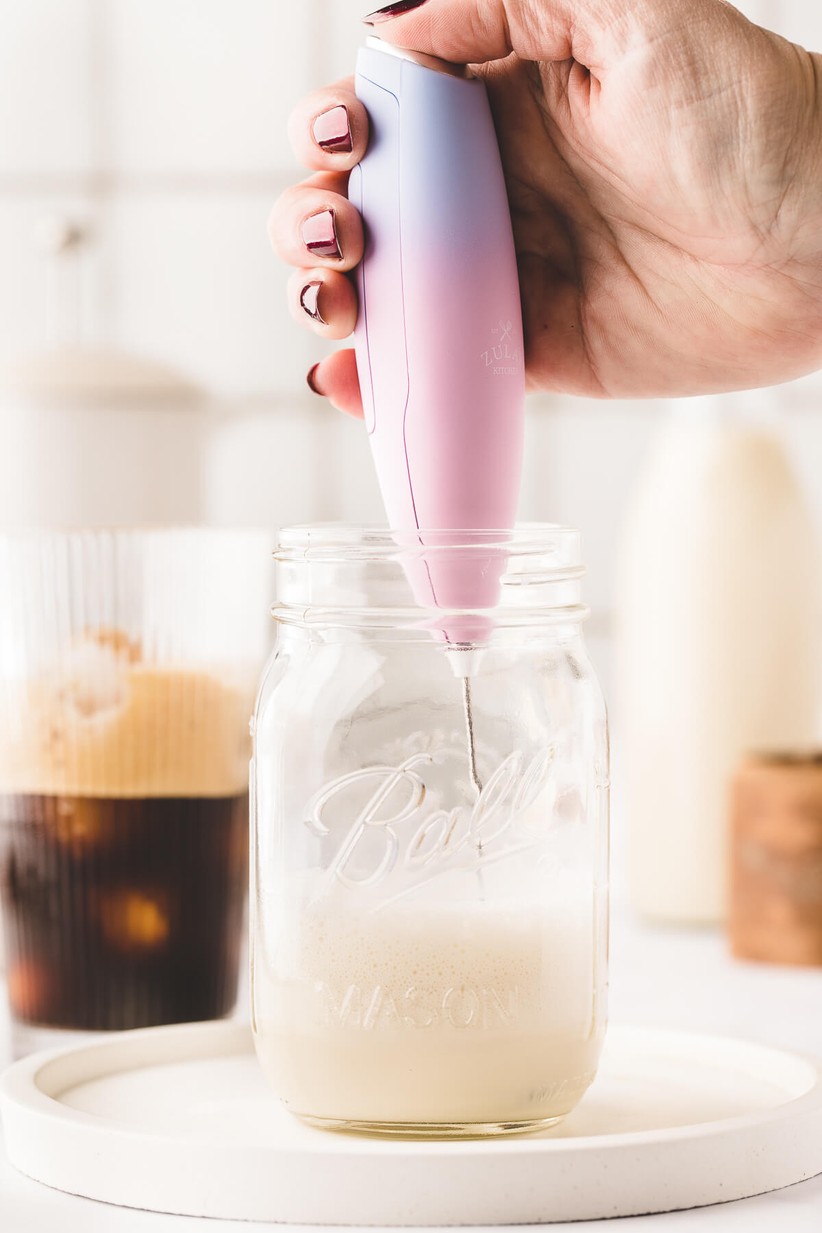 A pink milk frother makes cold foam out of oat milk in a mason jar. Shaken espresso in a glass can be seen in the background.