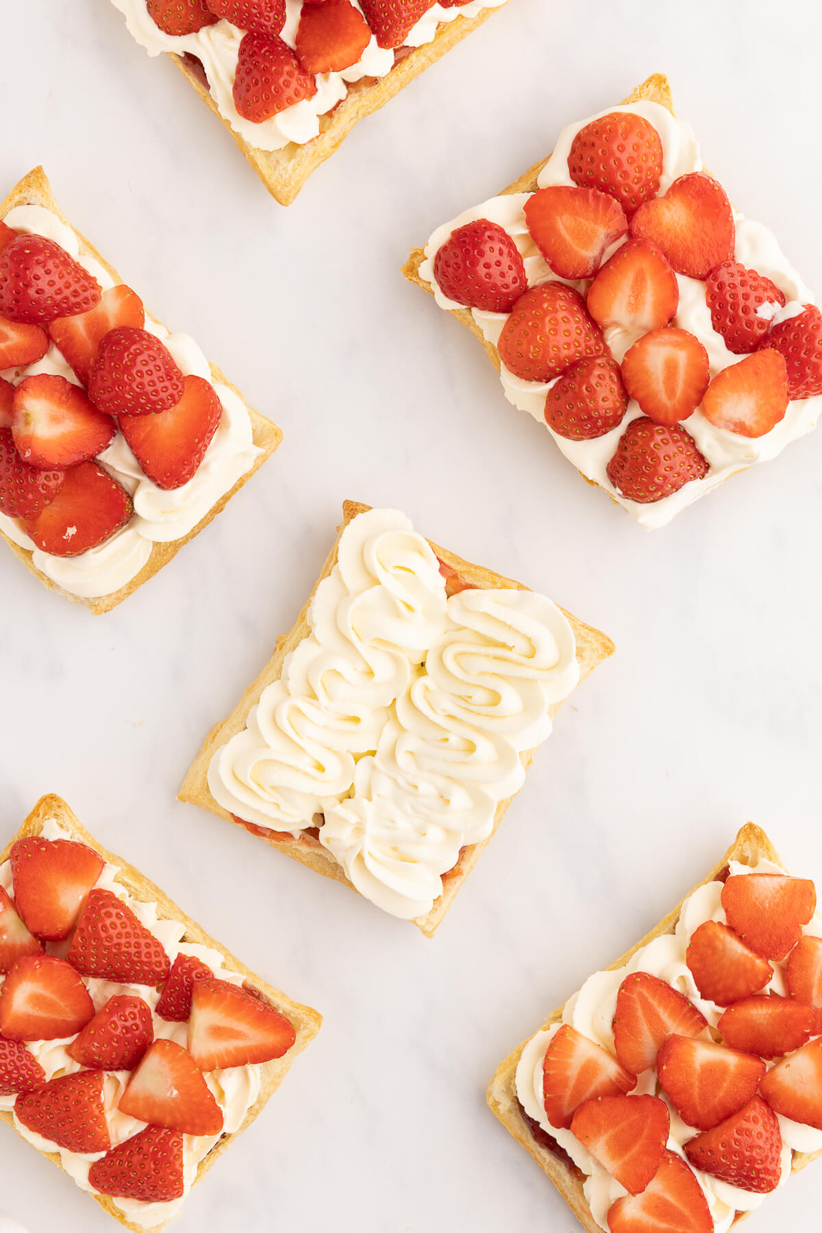 An overhead photo of how to make a Napoleon Dessert. 6 pieces of flaky puff pastry are topped with strawberry jam, swirls of whipped cream, and strawberries. This is how to make a delicious strawberry puff pastry Mille-Feuille. 
