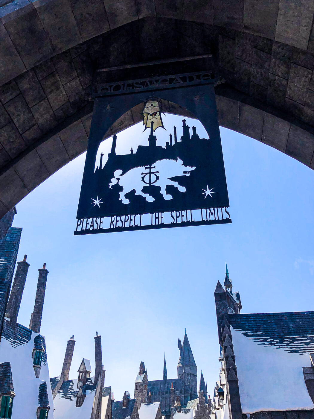 The stone entrance to The Wizarding World of Harry Potter™ at Universal Studios. A hogwarts sign says "please respect the spell limits." Snow capped building are seen in the background. 