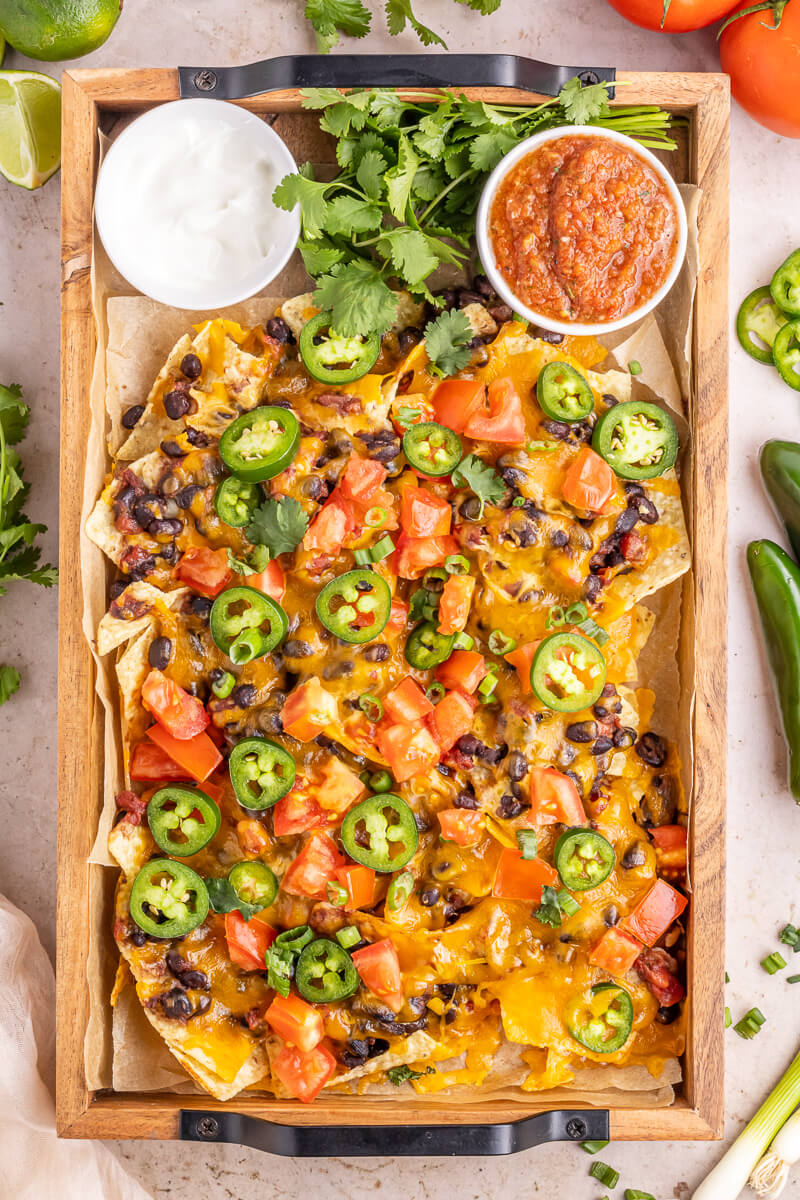 A wooden tray filled with vegetarian nachos with black beans and other toppings.