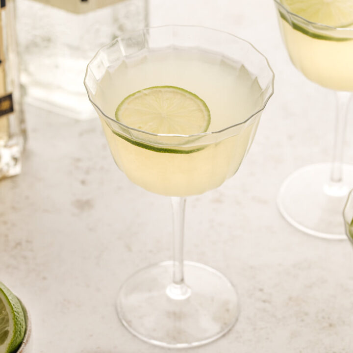 Three French gimlets with lime on a stone counter with a bottle of St. Germain in the background.