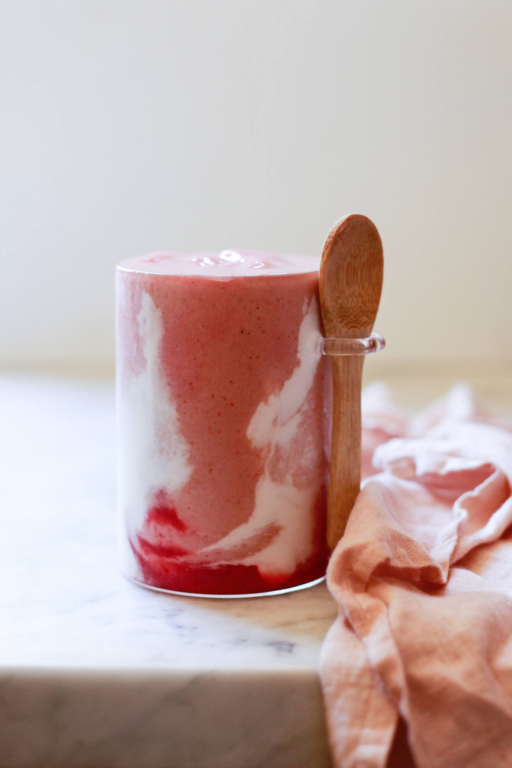 A beautiful copycat Hailey Bieber smoothie just like the Erewhon smoothie sits on a marble kitchen counter. The strawberry smoothie has red and white swirls and is in a jar with an attached wooden spoon. 