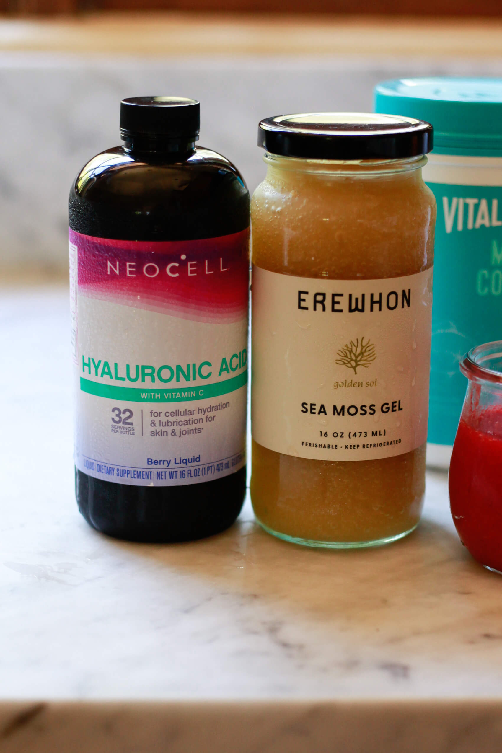 Neocell Hyaluronic acid liquid, Erewhon Sea Moss Gel, and Vital Proteins collagen sit on a marble kitchen counter. 